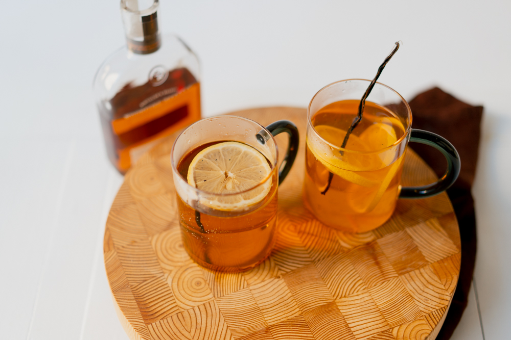 Two clean mugs filled with a light orange Hot Toddy cocktail, garnished with lemon slices and vanilla sticks. Placed upon a wooden cutting board with a bottle appearing out of focus in background. 