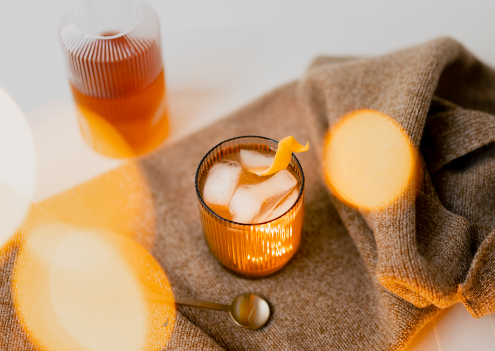 Gold coloured penicillin cocktail in a short glass, filled with ice and a garnish of lemon twist, set upon a brown cloth with a bottle of Scotch in the background 