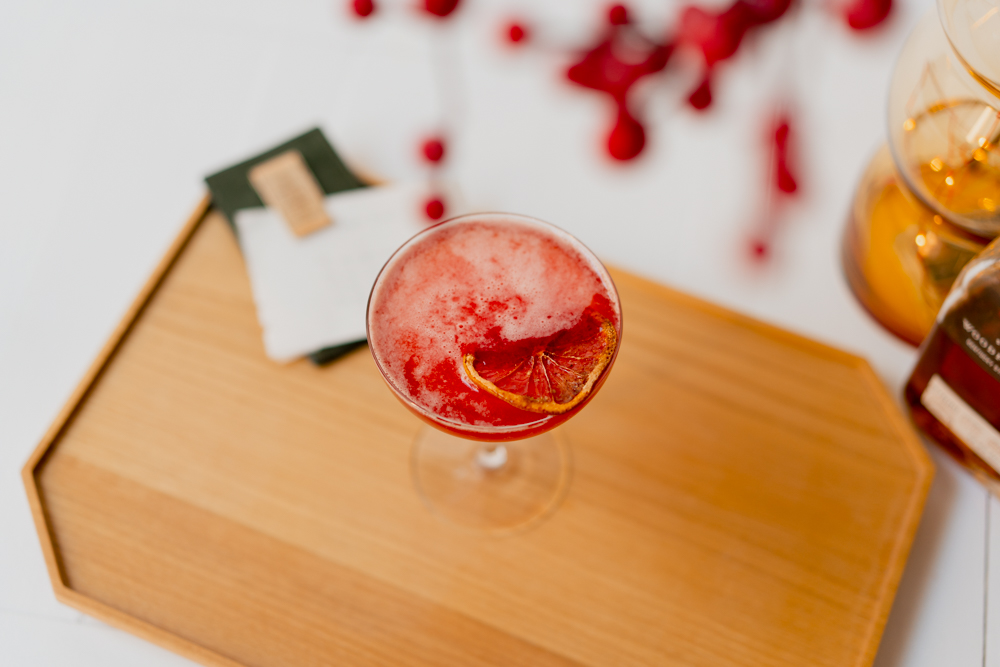 A red colour paper plane cocktail in a decorative glass, placed upon a wooden cutting board with a bottle of bourbon partially on screen 