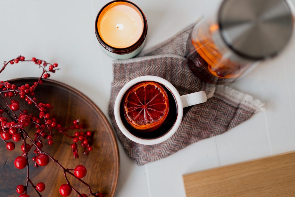 White coffee mug filled with red mulled wine and garnished with a slice of orange. Red holly berries, a lit candle and a glass container with the remaining beverage. 